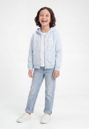 Παιδί με χαμόγελο και μαλλιά μέχρι τους ώμους, που φοράει μια ανοιχτό μπλε φούτερ με φερμουάρ και σχέδια, άσπρο μπλουζάκι, ανοιχτόχρωμο τζιν και άσπρα αθλητικά παπούτσια.