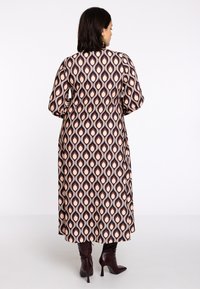 Langes Kleid mit einem abstrakten Muster in Braun, Creme und Pfirsich. Verfügt über puffte Ärmel und eine gerade Silhouette. Modell kombiniert mit burgunderfarbenen High Heels.