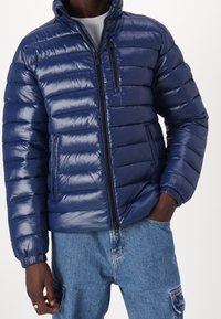 Marineblå dunjakke med vandret quiltet mønster, høj krave, lynlås foran og to sidelommer. Skinnende, glat tekstur.