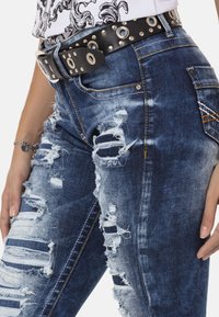 Blauwe versleten denim jeans met opvallende scheuren, met rafelige randen. Voorzien van een zwarte studs riem met metalen oogjes en een zilveren gesp.