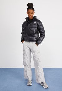 Mlada ženska z pletenicami, oblečena v črno puhasto jakno, bele cargo hlače, športne copate in črno torbico čez ramo, ki stoji na modri tla.