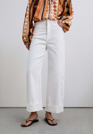 Persona vistiendo pantalones anchos blancos con puños, camisa de manga larga estampada metida por dentro, y sandalias planas negras con tachuelas, de pie sobre un suelo gris.