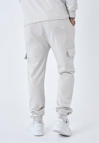 Lichtgrijze joggers van zacht materiaal, met zijzakken, elastische boorden en een subtiele branding op de achterkant. Gepaard met witte sneakers.
