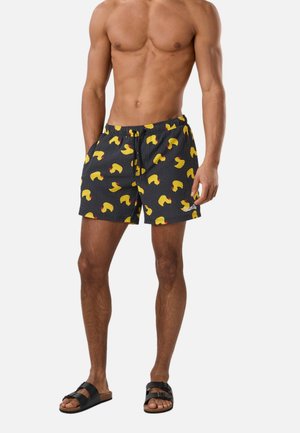 Mand uden skjorte iført sorte badebukser med gul andeprint og sorte sandaler, stående med afslappede hænder.