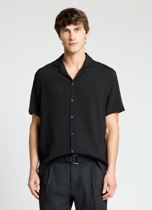 Mann mit heller Haut und braunen Haaren, der ein schwarzes kurzärmeliges Hemd mit Knöpfen und schwarze Hosen vor einem schlichten hellen Hintergrund trägt.