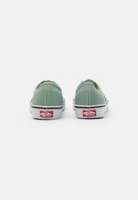 Grüne Canvas-Slipper mit weißen Gummisohlen, ausgestattet mit kontrastierenden Nähten und einem roten Logo an der Ferse.