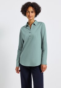 Camisetta verde chiaro a maniche lunghe con colletto, due bottoni alla chiusura e orlo curvo. Texture liscia, abbinata a pantaloni scuri.