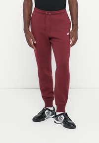 Burgundske trenirke z elastičnim pasom in vrvico; majhen bel Nike logotip na levem stegnu; v kombinaciji s črnimi športnimi copati, ki imajo srebrne detajle.