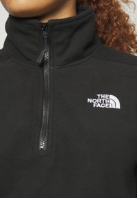 Chaqueta de forro polar negra con un cuello alto y cierre de media cremallera. Presenta un logo bordado blanco en el lado izquierdo del pecho y una textura suave.