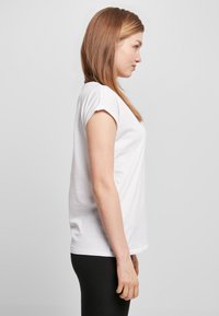 White cotton t-shirt with short rolled sleeves, relaxed fit, and crew neckline, paired with black leggings. Side view against a plain background.
