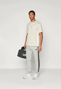 T-shirt beige Adidas con logo blu, pantaloni della tuta grigi con strisce nere e sneakers bianche. Tiene una borsa da palestra Adidas nera.