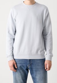 Homme portant un sweat-shirt gris clair uni à col rond et un jean bleu, debout devant un fond blanc.
