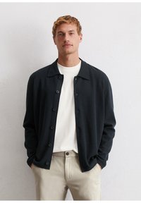 Dark navy textured cardigan with a collar, button closure, worn over a white shirt, paired with light-colored pants.