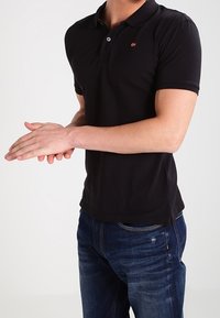 Svart polo skjorta med en liten logotyp på bröstet, i kombination med blå denimjeans. Skjortan har korta ärmar och en klassisk krage.