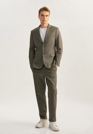 Traje oliva entallado con americana de un solo botón, solapas en pico, dos botones, pantalones a juego y una camiseta blanca debajo. Zapatillas blancas informales.