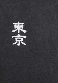 Černé tričko vyrobené z látky s bílými japonskými znaky. Materiál má jemnou texturu a hladký povrch. Design se zaměřuje na výrazný grafický text.