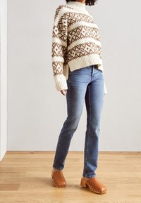 Suéter tejido grueso con patrón blanco y marrón, jeans azul de pierna recta, y zuecos marrones con tachuelas plateadas, sobre un fondo neutro.