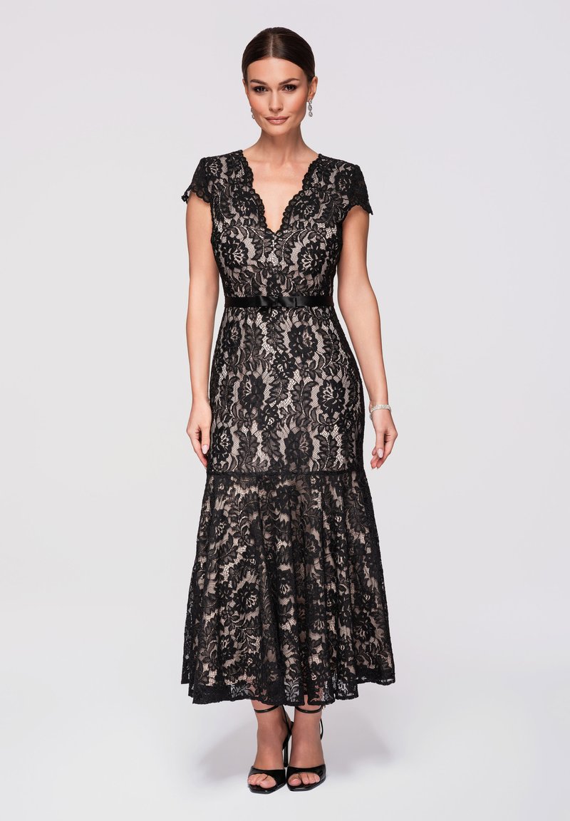 Femme portant une robe noire en dentelle à col en V ajustée avec manches courtes, ceinture en satin et ourlet évasé, assortie à des sandales noires à talons hauts.