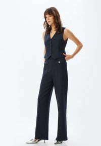 Marinblå randig väst med V-ringning och knäppning fram, ihop med vida marinblå byxor med knappdetaljer. Silverfärgade spetsiga skor.