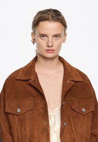 Giacca di suede marrone con colletto, due tasche frontali e bottoni a pressione argentati, abbinata a una maglietta color crema con rifiniture in pizzo.