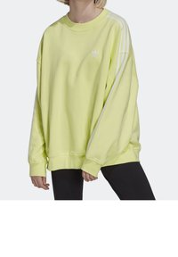 Světle žlutá oversized mikina s kulatým výstřihem, zdobená bílými třemi pruhy na ramenou a malým logem na prsou.