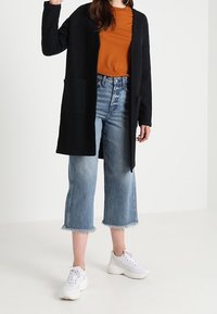 Musta neuletakki avoimella edustalla ja kahdella taskulla, päällä oranssi t-paita. Rento vaaleansininen lyhytfarkku ja valkoiset lenkkarit täydentävät asun.