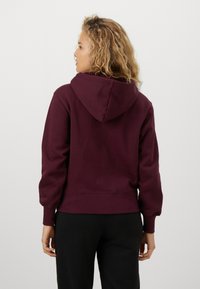 Bordeauxfärgad hoodie med dragsko i huvan, ribbade muddar och nederkant, samt en mjuk textur. Visad från baksidan, tillsammans med svarta byxor.