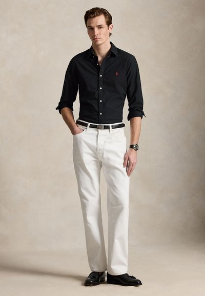Ung mand iført en sort skjorte med knapper, hvide bukser med sort bælte, sorte loafers og et armbåndsur, stående med den ene hånd i lommen.