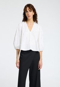 Blouse blanche raccourcie avec des manches bouffantes et un col en V, présentant de subtils détails brodés dans un design abstrait. Assortie à un pantalon large noir.