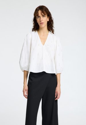 Blouse blanche raccourcie avec des manches bouffantes et un col en V, présentant de subtils détails brodés dans un design abstrait. Assortie à un pantalon large noir.