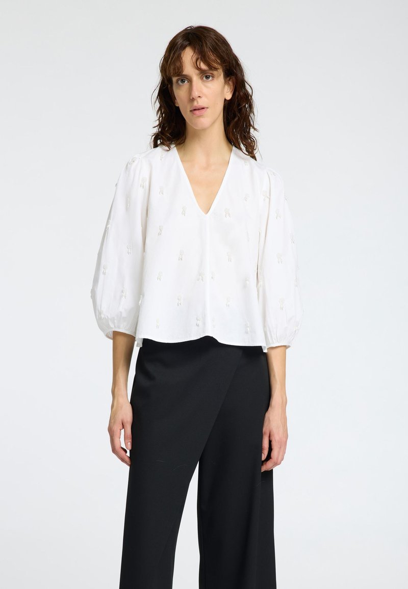 Blouse blanche raccourcie avec des manches bouffantes et un col en V, présentant de subtils détails brodés dans un design abstrait. Assortie à un pantalon large noir.