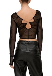 Schwarzes, transparenter Langarm-Top mit gerafftem Design, das einen Schlüssel-Ausschnitt und ein Bindeband im Rücken hat, kombiniert mit hochgeschnittenen schwarzen Hosen.