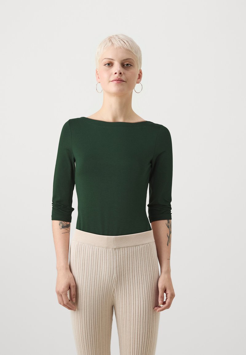 Donkergroene blouse met lange mouwen en een bootnek ontwerp, gecombineerd met beige geribbelde hoge taille broeken. Zachte textuur en strakke pasvorm.
