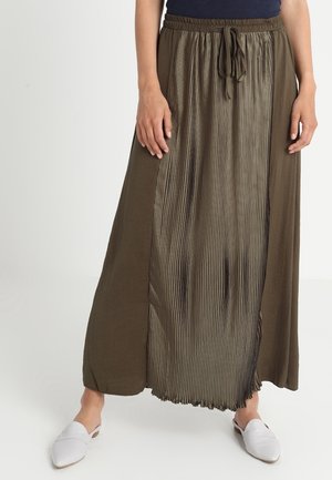 Kvinde iført en lang olivengrøn nederdel med et plisseret panel og snøre i taljen, kombineret med hvide slip-on sko.