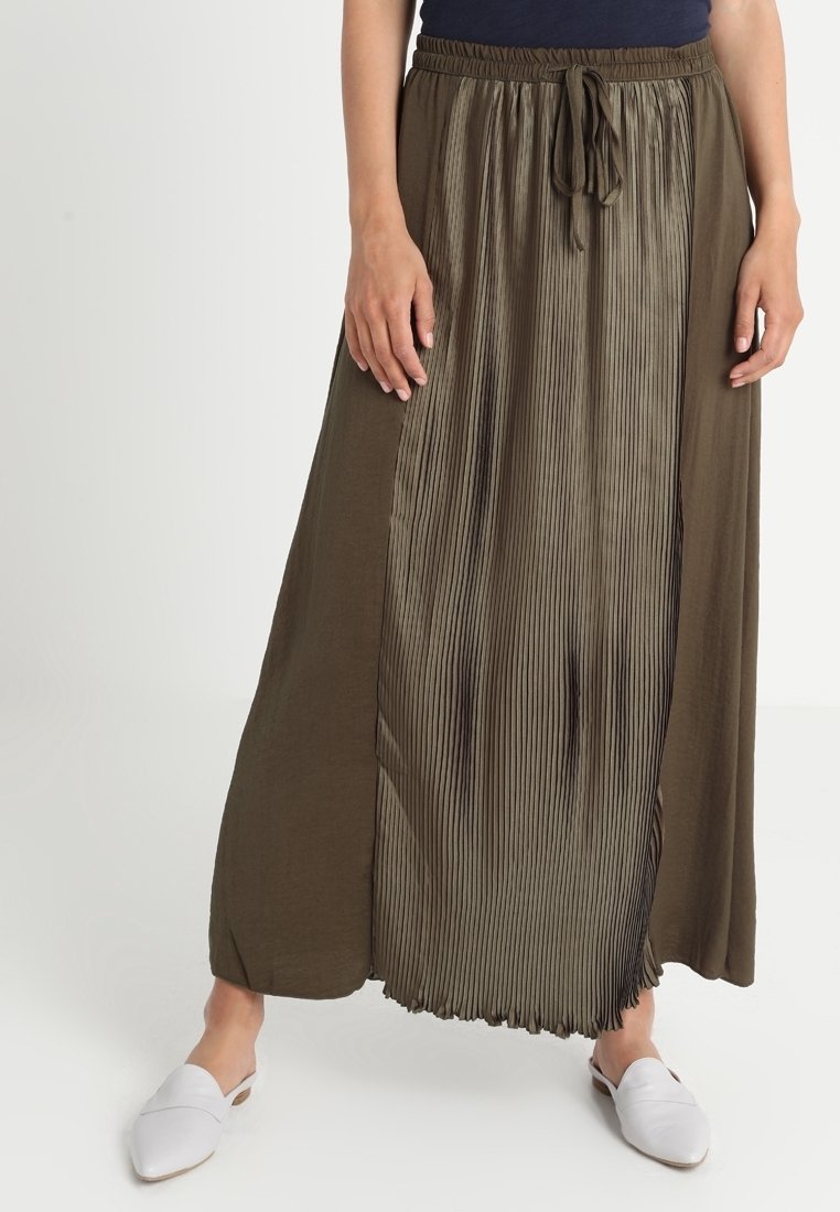 Femme portant une longue jupe vert olive avec un panneau plissé et une taille à cordon, associée à des chaussures blanches slip-on.