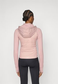 Párnázott rózsaszín kabát puha textúrával, testhezálló kialakítással és kapucnival. Hosszú ujjakkal és a hát alsó részén logóval rendelkezik. Fekete leggingszel viselve.