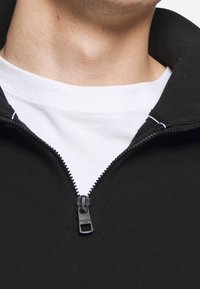 Man in een witte crewneck-shirt onder een zwarte zip-up trui met de ritssluiting gedeeltelijk dicht bij de nek.