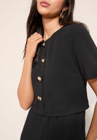 Fekete rövid ujjú felső kerek nyakkivágással, arany díszgombokkal az elején és sima anyagtextúrával.