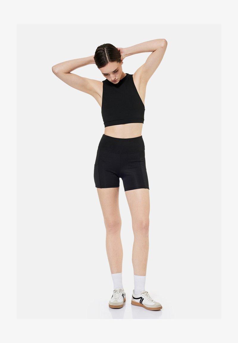 Joven mujer con crop top negro sin mangas y pantalones cortos de cintura alta, arreglándose el pelo, lleva calcetines blancos y zapatillas deportivas casuales.