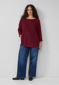 Bordeauxfarbenes Oversized-Oberteil mit rundem Ausschnitt und 3/4-Ärmeln, kombiniert mit weit geschnittenen blauen Denim-Jeans und schwarzen Knöchelstiefeln.