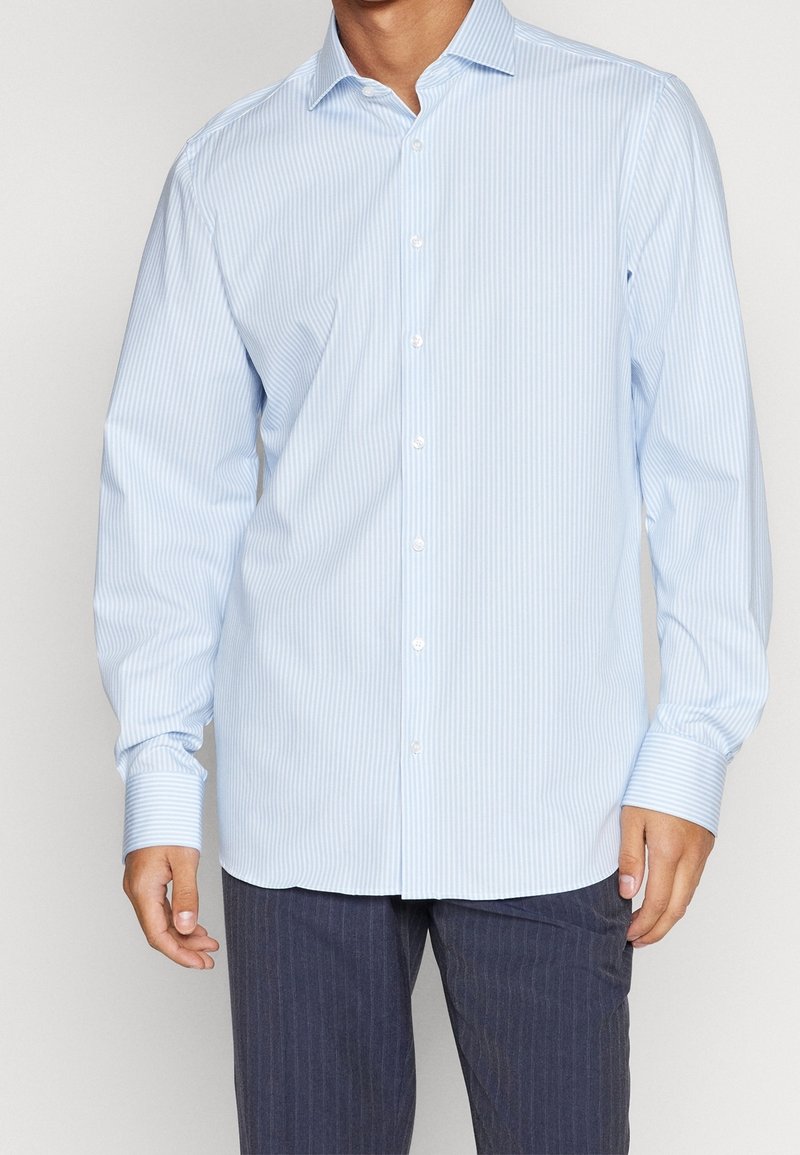 Homme portant une chemise bleu clair à rayures verticales et un pantalon bleu foncé à fines rayures, montré des épaules aux hanches.