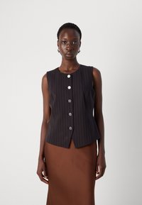 Gilet senza maniche a righe navy con scollo rotondo, caratterizzato da cinque bottoni rotondi argento e una vestibilità sartoriale. Una gonna marrone completa il top.