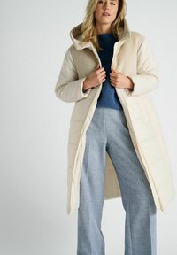 Beige pufferjack met een zachte capuchon, rits aan de voorkant en gewatteerde textuur, gelaagd over een blauwe trui en grijze wijde broek.