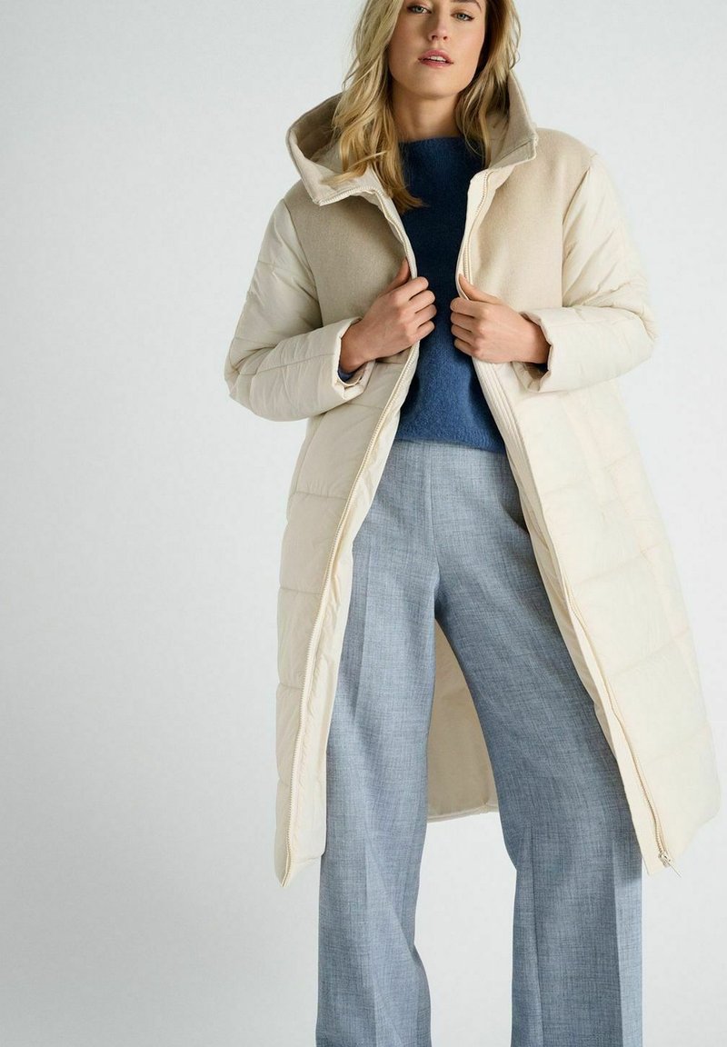 Beige pufferjack met een zachte capuchon, rits aan de voorkant en gewatteerde textuur, gelaagd over een blauwe trui en grijze wijde broek.