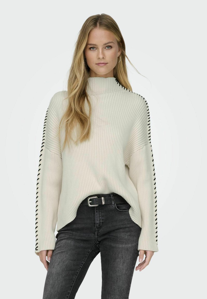 Maglione a collo alto a coste crema con dettagli a striscia nera sui lati, abbinato a jeans scuri e una cintura nera. Tessuto morbido, vestibilità rilassata.