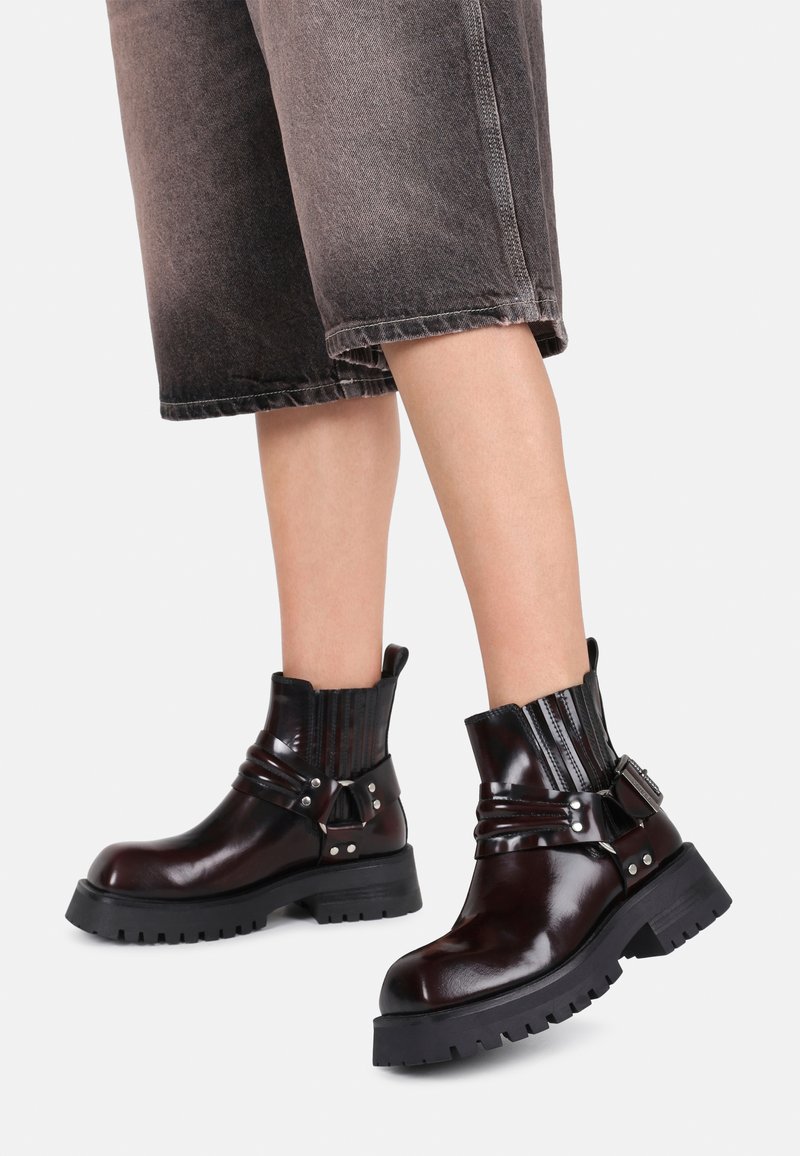Bottines en cuir marron avec une semelle noire épaisse, dotées de sangles, de détails en métal argenté et d'un tige côtelée. Les bottines offrent un design robuste.