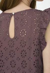 Top viola con motivo a fiori in pizzo, maniche a volant e chiusura con piccolo bottone sul retro. Presenta un'apertura a goccia sul retro.