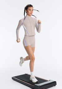 Femme courant sur un tapis de course noir portant une veste de sport beige zippée et un short avec des rayures blanches et bleues, ainsi que des baskets blanches.