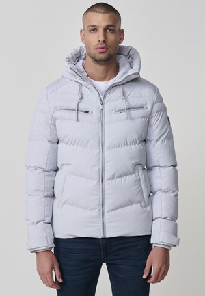 Chaqueta acolchada gris claro con cierre frontal, dos bolsillos en el pecho con cremallera y capucha. Presenta paneles horizontales cosidos y puños elásticos.
