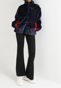 Marinblå fuskpälsjacka med hög krage, röda fickor och dragkedja framtill, parat med svarta utsvängda byxor och vita sneakers.